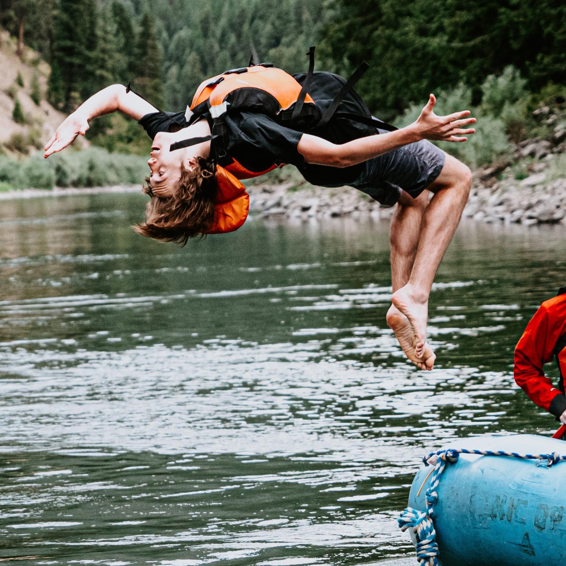 Person performing a backflip off a blue raft into the Clarkfork River, while another person sits on the raft holding a paddle, with a second raft and tree-lined shore in the background