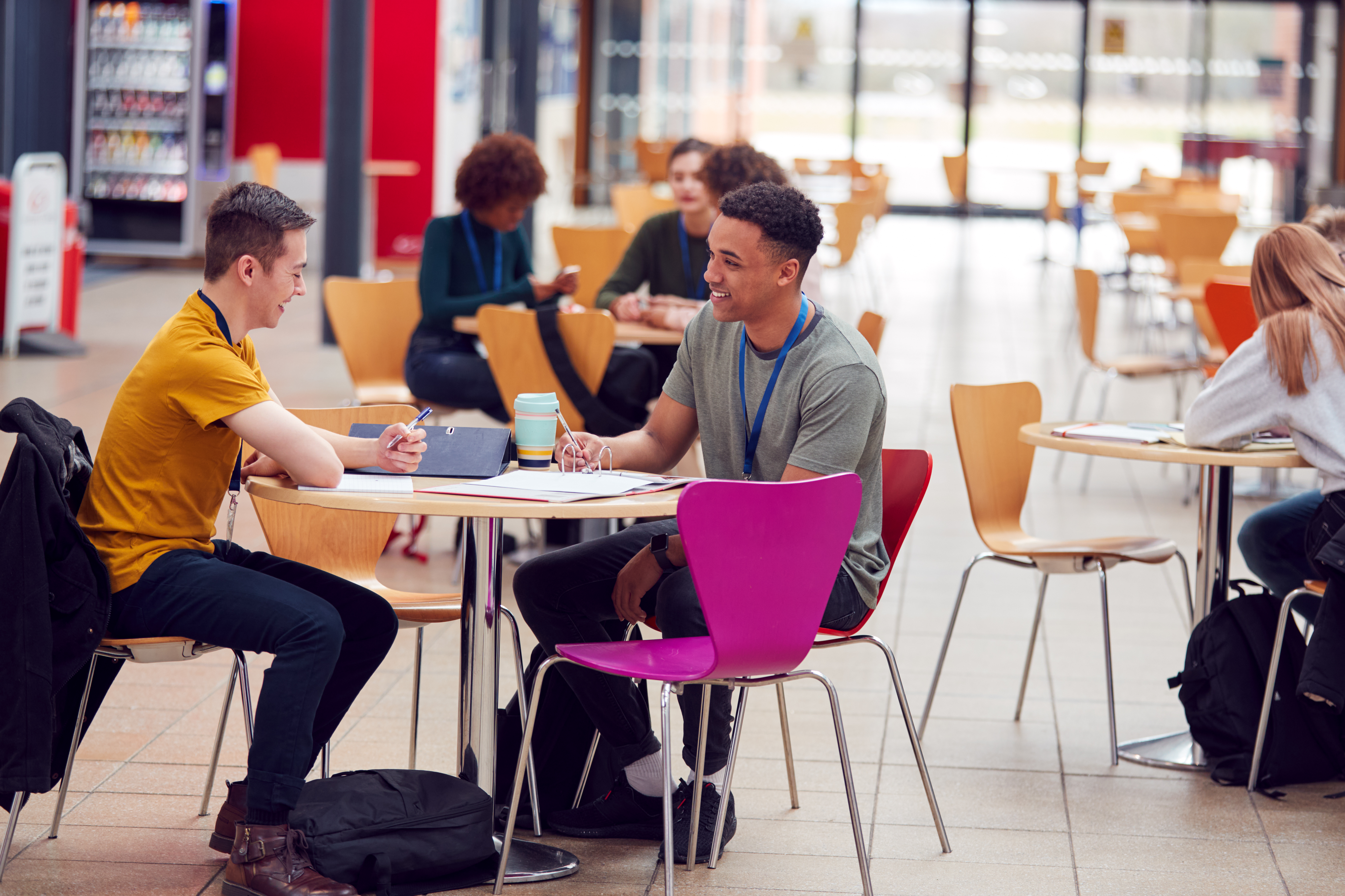 Two young men engaged in conversation at a wooden table in a brightly lit university cafeteria, surrounded by other students