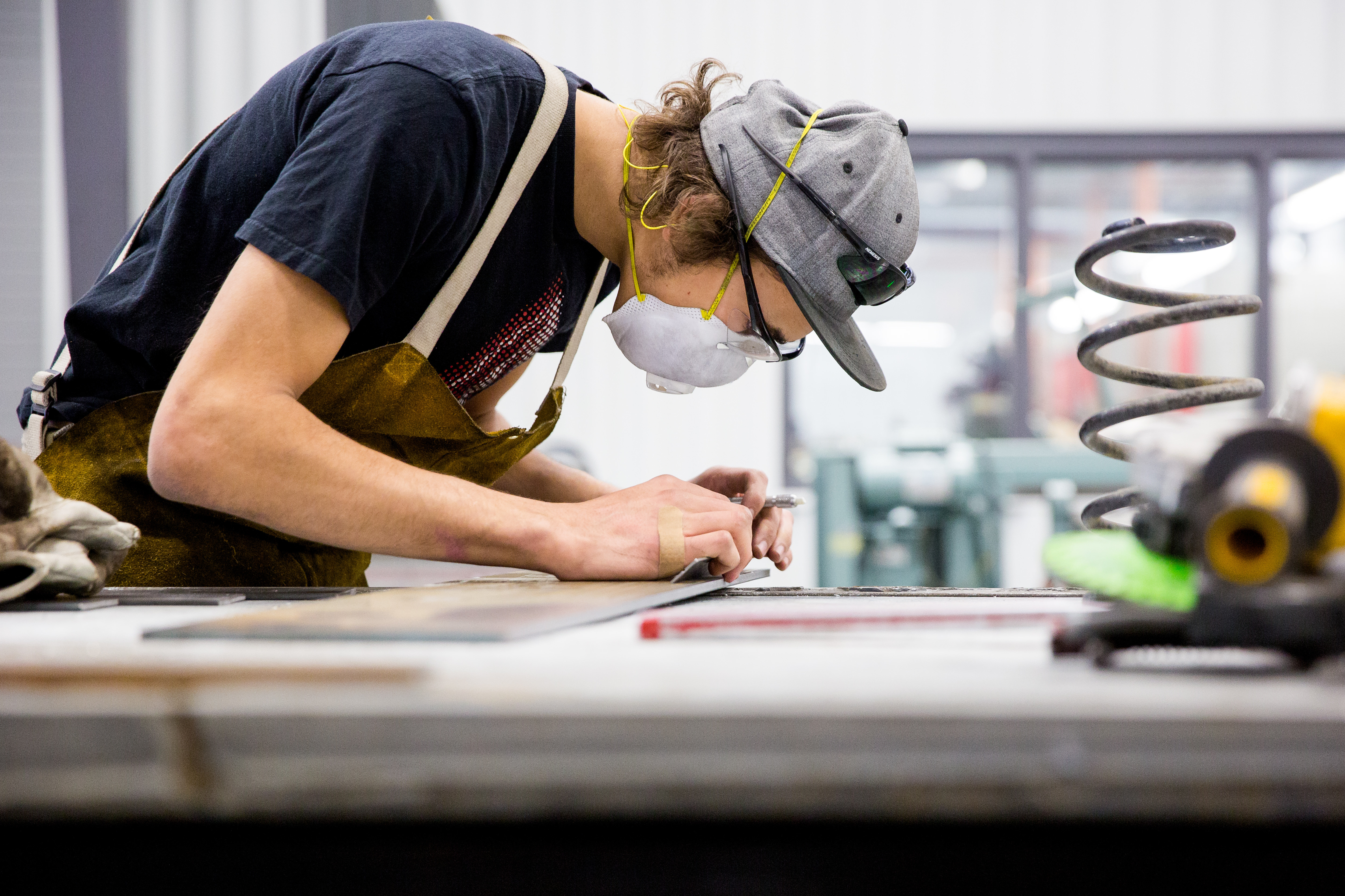 shows a person working in a workshop or industrial setting. They are wearing protective gear, including a face mask, safety glasses, and ear protection, along with an apron.