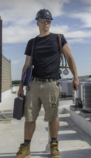 A technician standing on a rooftop, wearing a hard hat and safety glasses, holding a toolbox and equipment. HVAC units are visible behind him, and he has gauges and tools clipped to his belt and shoulder straps